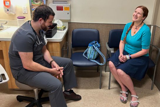 A man with dark hair and beard, in gray scrubs, talks with a woman with long braided hair and blue top in a clinic exam room.